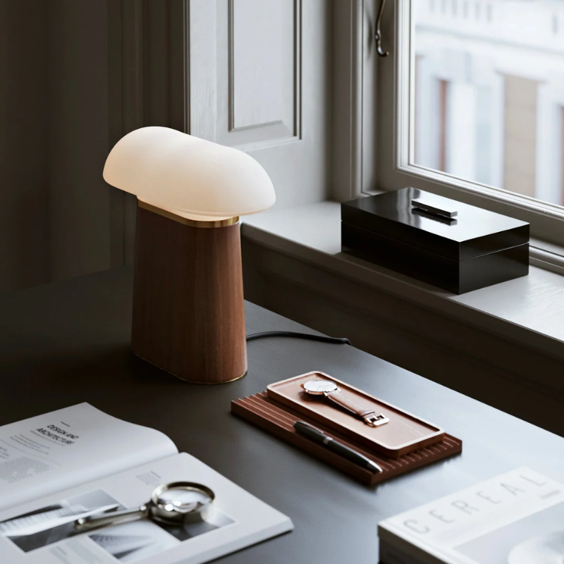 The Nova Table Lamp by Woud in walnut with an opal glass shade on a desk.